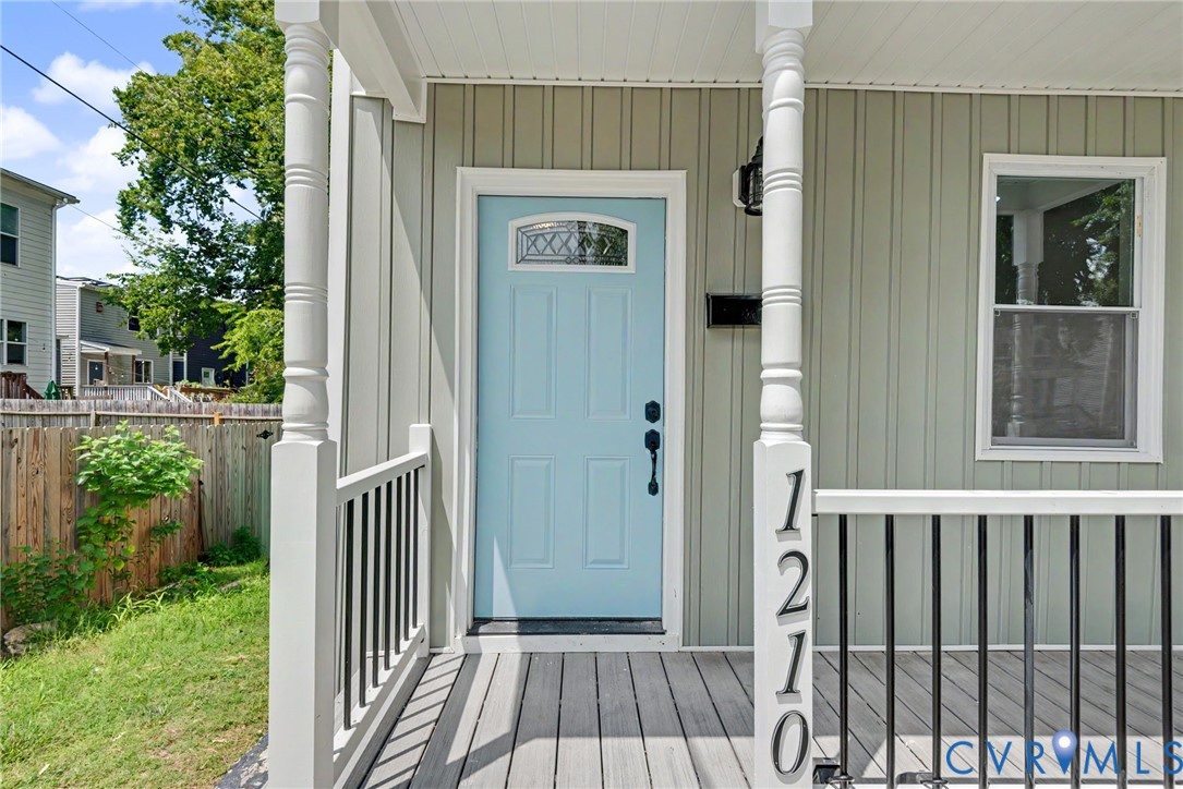 Covered front porch featuring board-and-batten siding, a light blue entry door with decorative glass inlay, turned porch columns, and a black metal railing