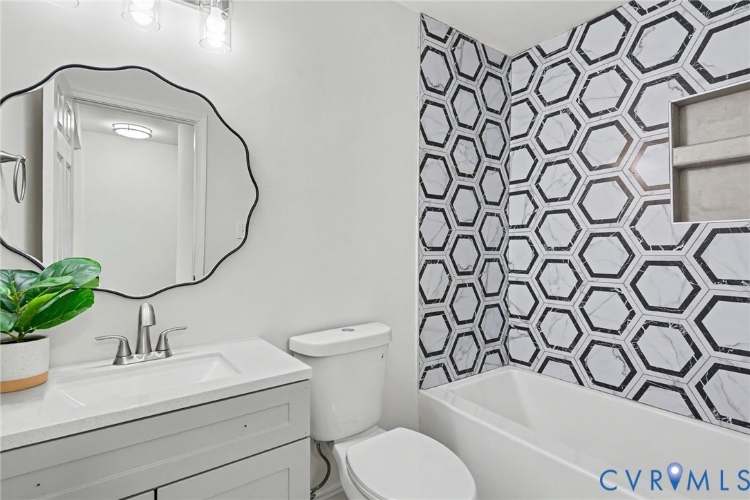 Grey vanity with integrated countertop and sink, scalloped-edge mirror, multi-light fixture, and bathtub featuring a hexagonal patterned tile surround with a built-in shower niche