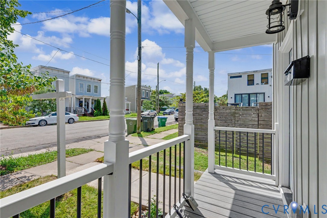 Covered front porch featuring white structural columns, black metal railings, wood-finish flooring, and a wall-mounted exterior light fixture