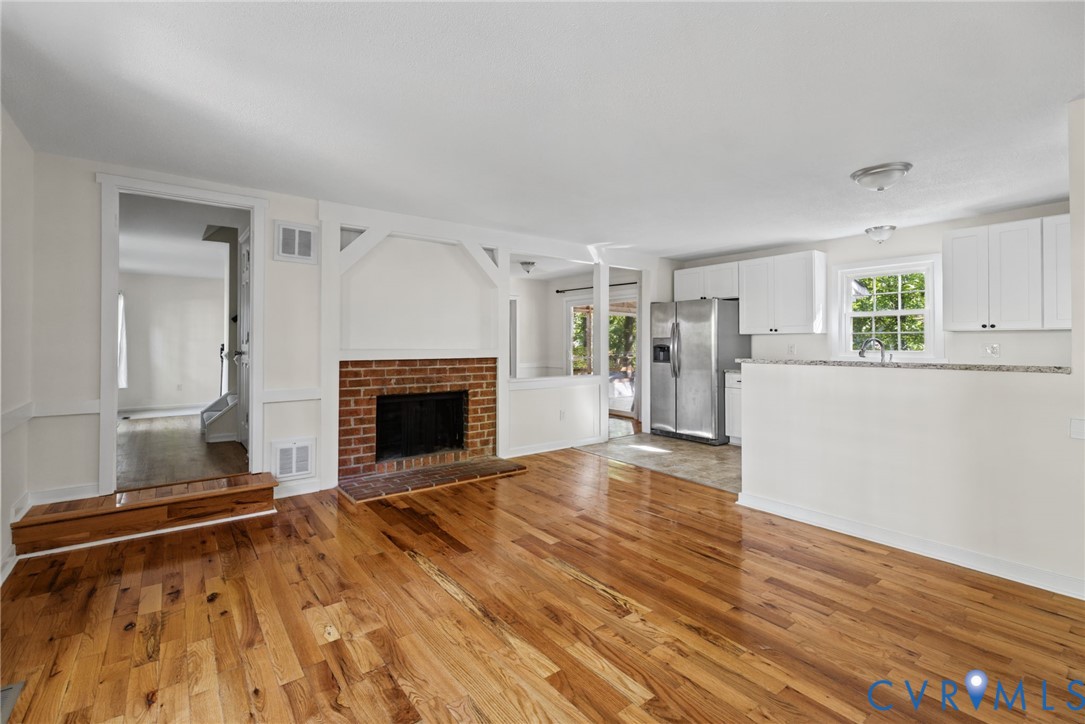 Wood-finish flooring, brick fireplace with white decorative framing, kitchen with stone-surface countertops, white cabinetry, and stainless steel refrigerator
