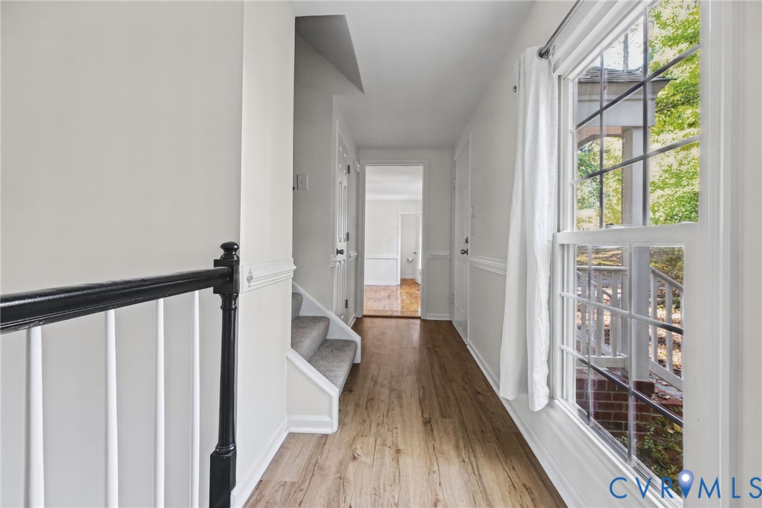 Hallway featuring wood-finish flooring, chair rail molding, a staircase with a black handrail, and windows with multi-pane grilles Hallway featuring wood-finish flooring, chair rail molding, a staircase with a black handrail, and windows with multi-pane grilles