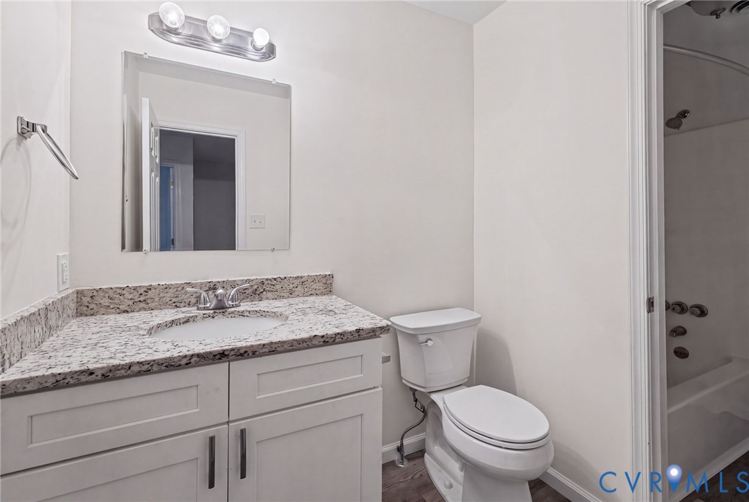 Bathroom featuring white cabinetry, stone-surface countertop with integrated basin, frameless wall-mounted mirror, three-light vanity fixture, and wood-finish flooring