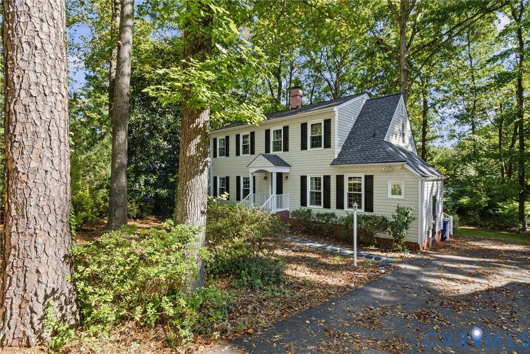 Two-story colonial-style residence featuring light-toned horizontal siding, dark decorative shutters, a gabled front entryway, a steep-pitched roof, and mature perimeter trees
