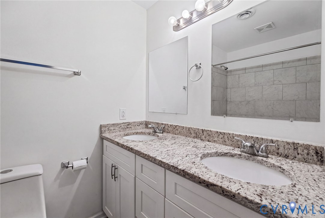 Dual-sink vanity with a stone countertop, white shaker-style cabinetry, chrome fixtures, and a tiled shower surround