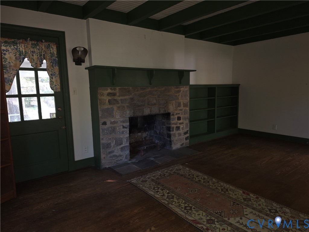 Stone fireplace with a mantle, built-in shelving, exposed ceiling beams, wood-finish flooring, and a multi-pane glass door
