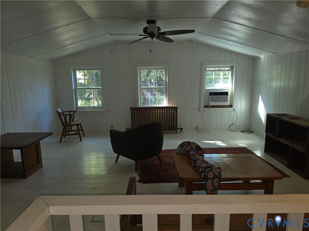 Vaulted ceiling with vertical wood-paneled walls, multiple windows, metal radiator, and ceiling fan