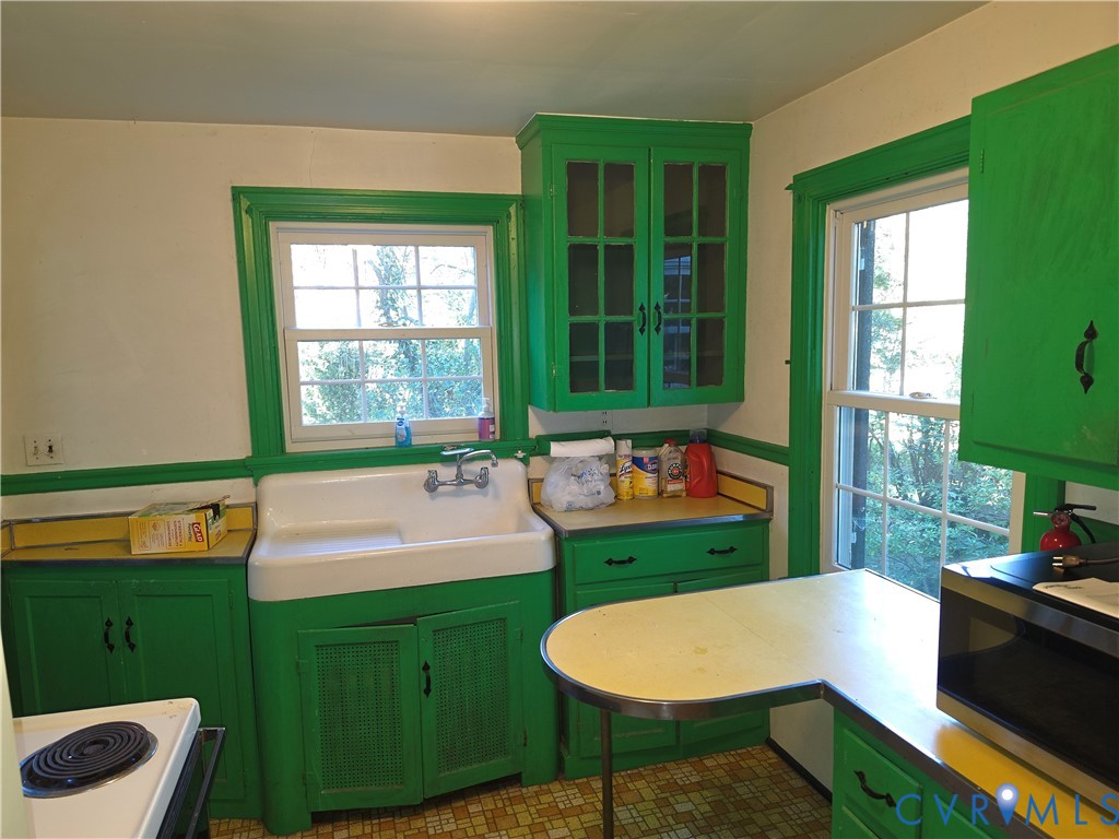 Kitchen featuring a farmhouse-style apron sink, custom cabinetry with glass-front uppers, solid-surface countertops with integrated metal edging, and dual windows