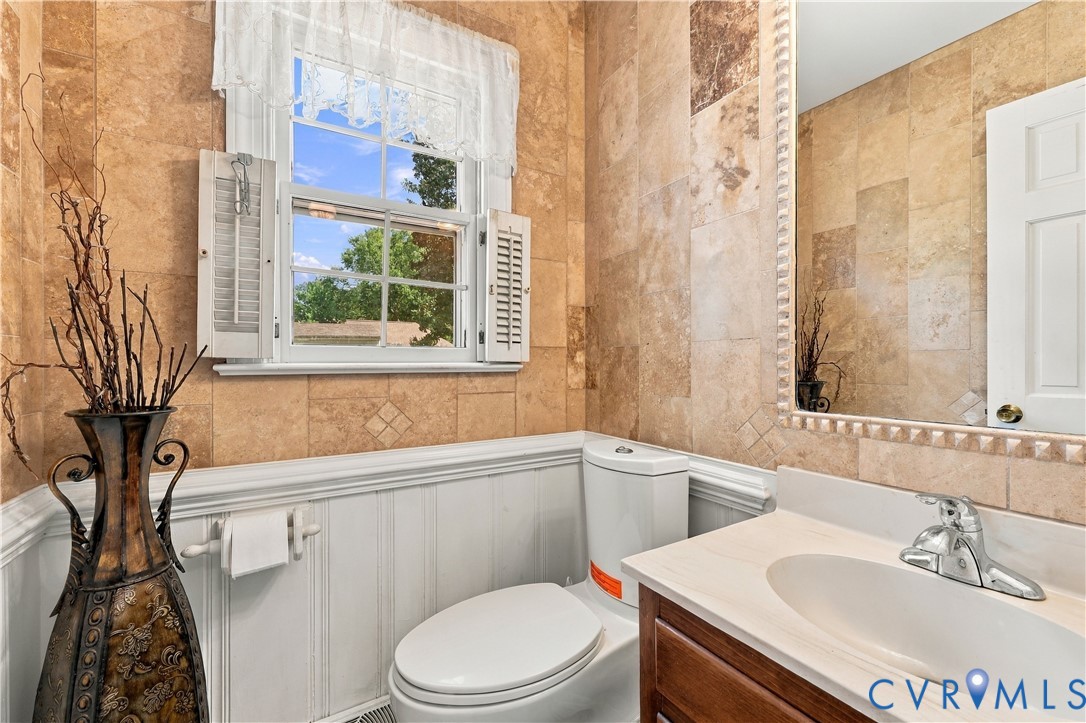Full-wall textured tile backsplash, vanity cabinet with inset sink, polished chrome faucet, white wainscoting, and wood-framed window with integrated shutters