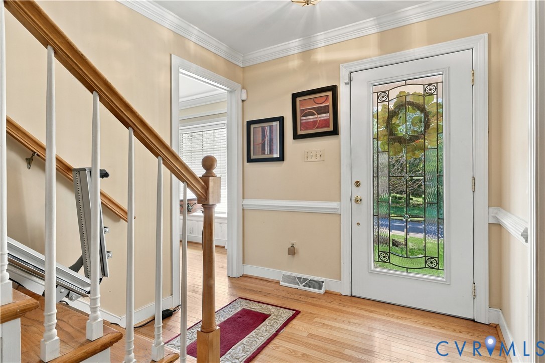 Entryway featuring wood-finish flooring, a white front door with an intricate decorative glass panel, crown molding, chair rail trim, and a wooden staircase railing with white balusters