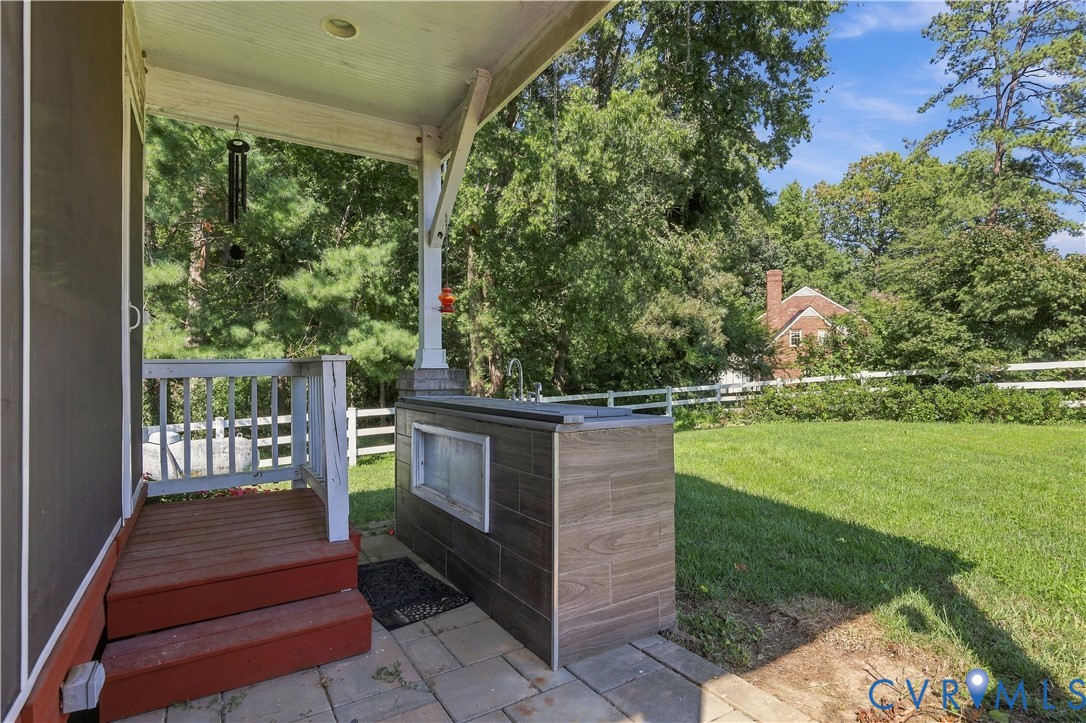Covered patio featuring paver flooring, built-in outdoor kitchen with wood-finish tile cladding, and wood-railing stairs, overlooking a manicured lawn enclosed by a white fence
