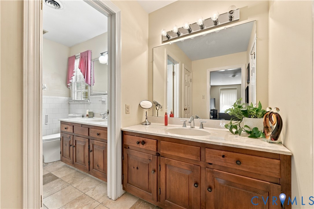 Bathroom featuring wood-finish cabinetry, neutral stone-finish countertops, undermount sinks, full-width vanity mirrors, and multi-bulb strip lighting