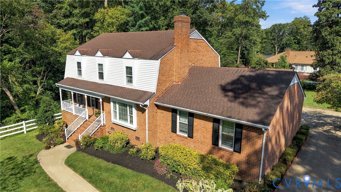 Dutch Colonial exterior featuring red brick masonry, white vinyl siding, a multi-gabled roofline, a prominent brick chimney, and a covered front porch with white railing