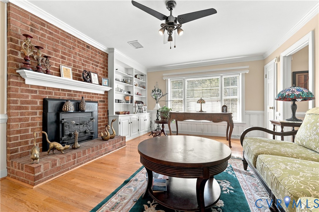 Living area featuring a floor-to-ceiling brick fireplace with a mantle, built-in shelving, wood-finish flooring, crown molding, and a bay window