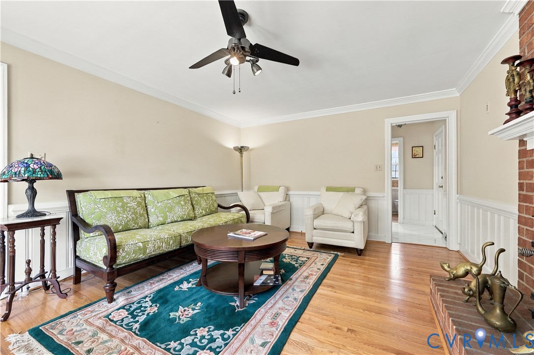 Living area featuring wood-finish flooring, brick fireplace hearth, crown molding, chair rail wainscoting, and a ceiling fan with integrated lighting