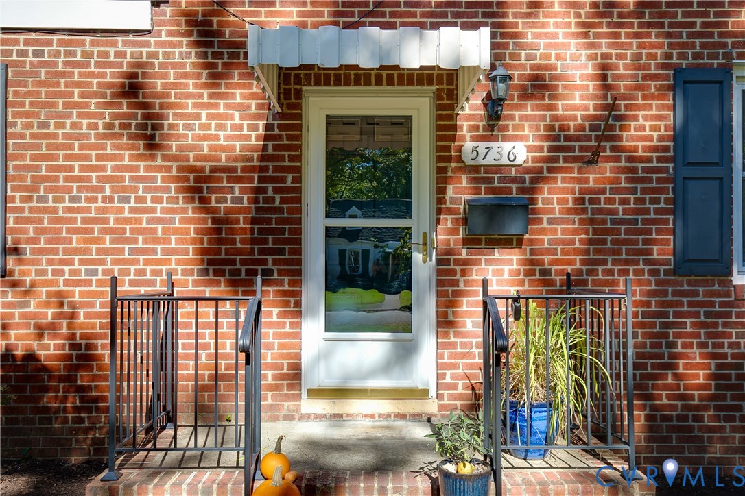 Red brick exterior featuring a white storm door, scalloped metal awning, dark-toned shutters, and a black wall-mounted mailbox Red brick exterior featuring a white storm door, scalloped metal awning, dark-toned shutters, and a black wall-mounted mailbox