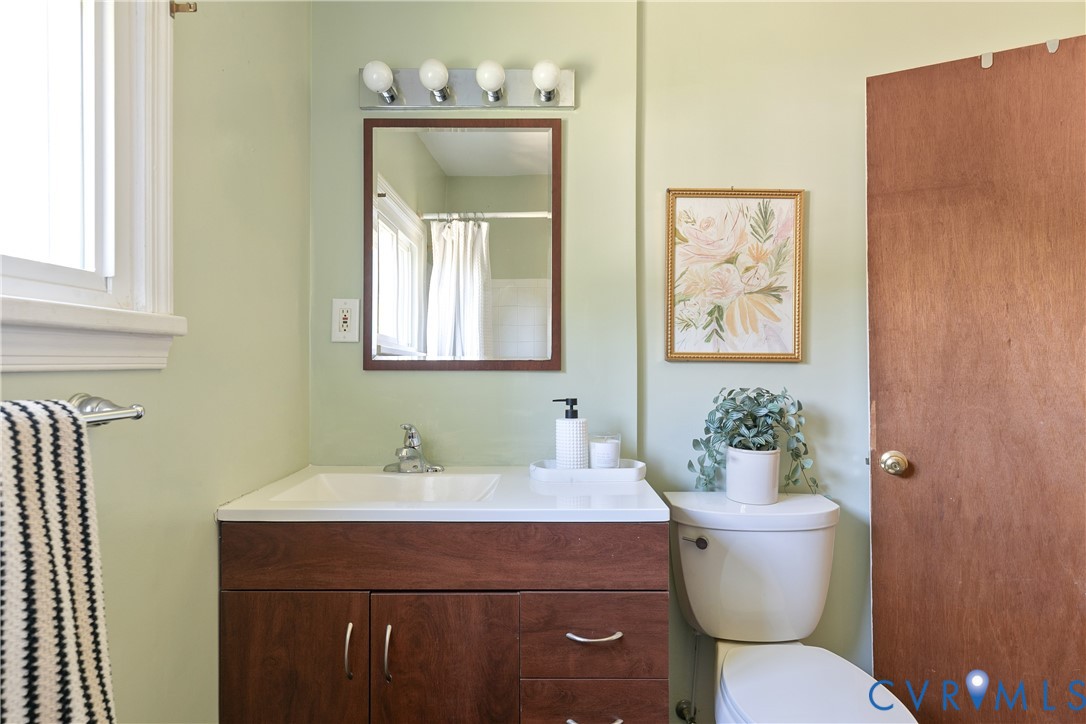 Bathroom featuring a wood-finish vanity with integrated sink, rectangular wall-mounted mirror, four-bulb vanity light, and white porcelain toilet