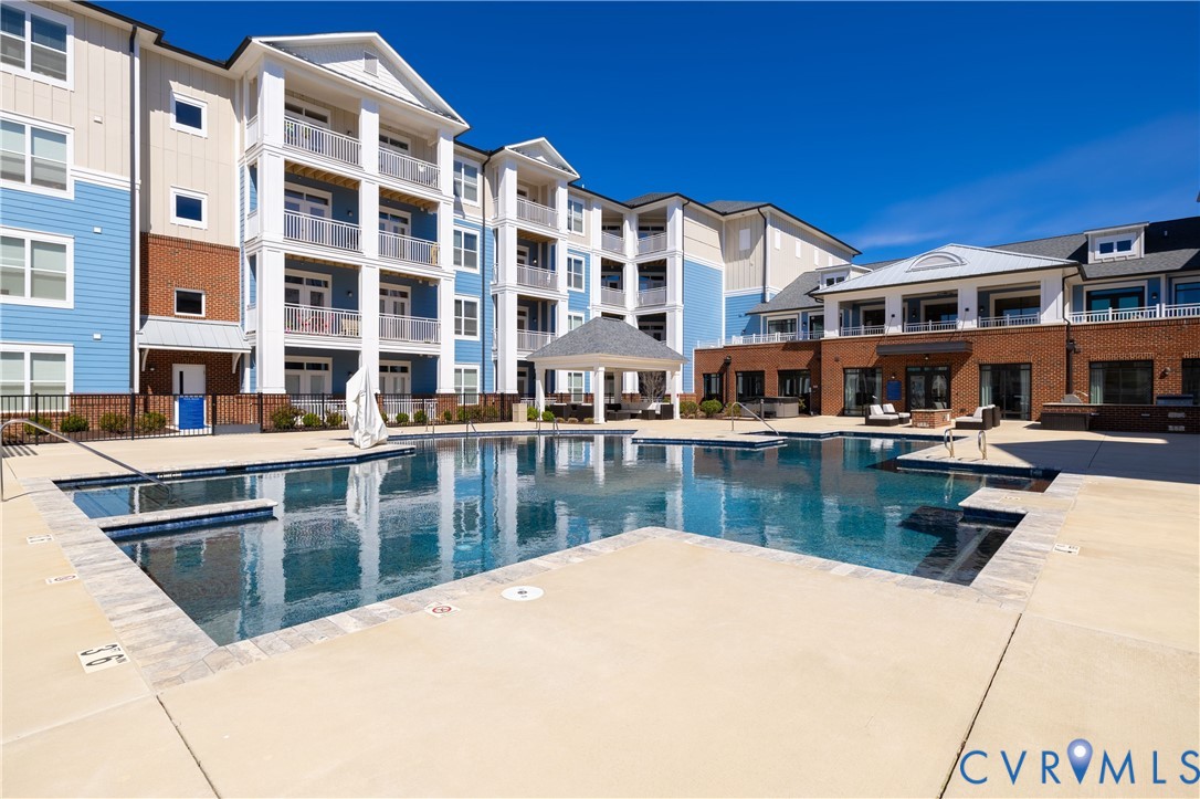 Swimming pool featuring a paved deck, perimeter fencing, covered pavilion, and multi-story building exterior with brick and siding finishes