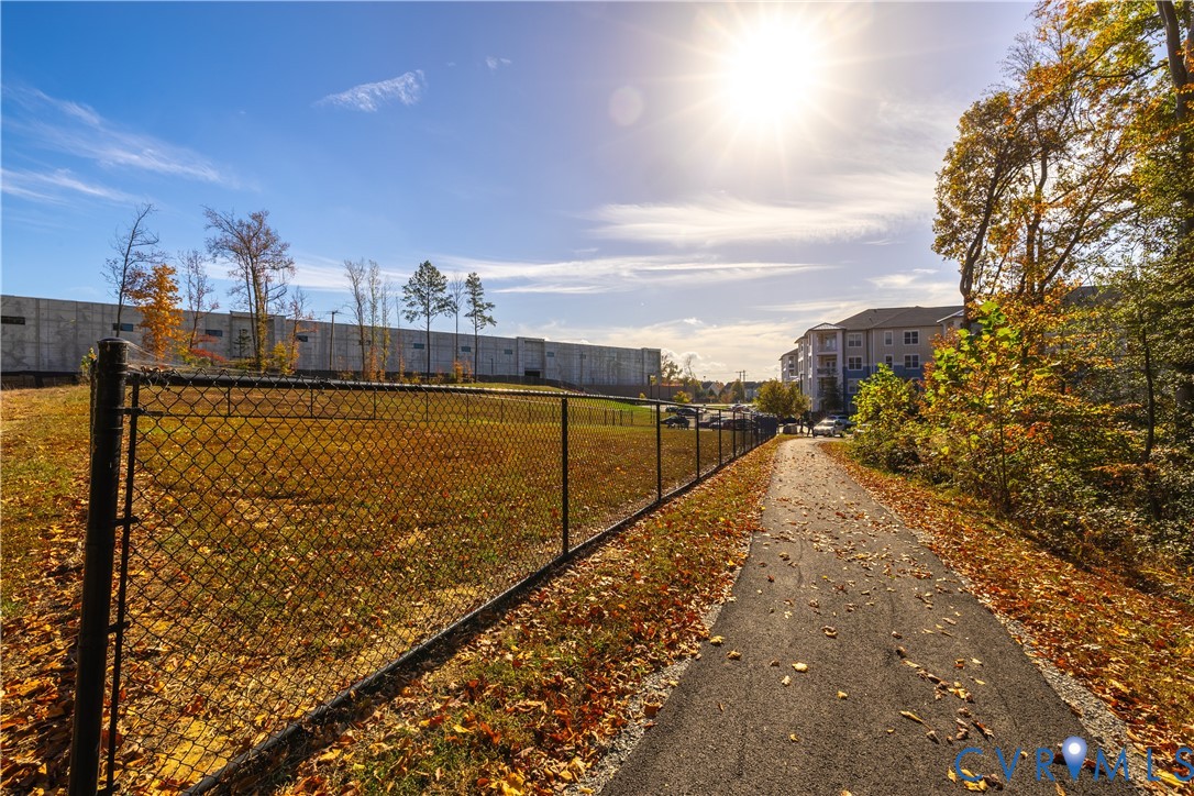 Paved trail bordering an open grass lot enclosed by a chain-link fence, surrounded by mature trees