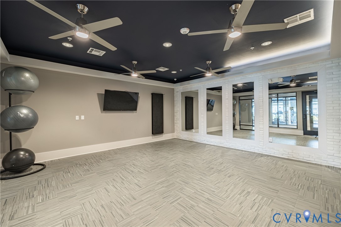Spacious room featuring a dark-toned tray ceiling with recessed lighting, four industrial-style ceiling fans, wall-to-wall mirrors, textured white brick accent wall, and patterned tile flooring