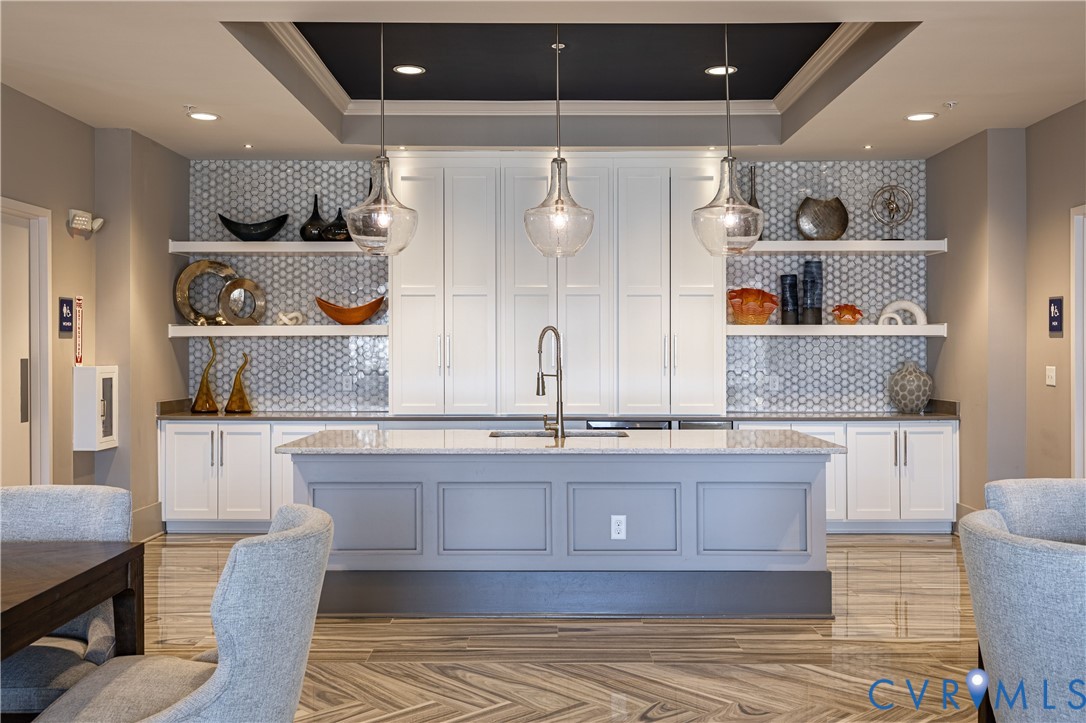 Central island with stone countertop and undermount sink, white cabinetry with recessed-panel doors, hexagonal tile backsplash, floating wall shelving, and tray ceiling with integrated pendant lighting Central island with stone countertop and undermount sink, white cabinetry with recessed-panel doors, hexagonal tile backsplash, floating wall shelving, and tray ceiling with integrated pendant lighting