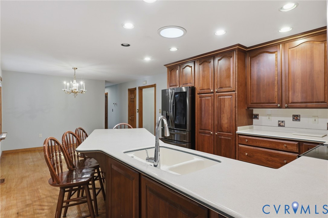 Kitchen featuring rich wood-finish cabinetry, a central island with a dual-basin sink, high-reach faucet, neutral solid-surface countertops, and recessed lighting