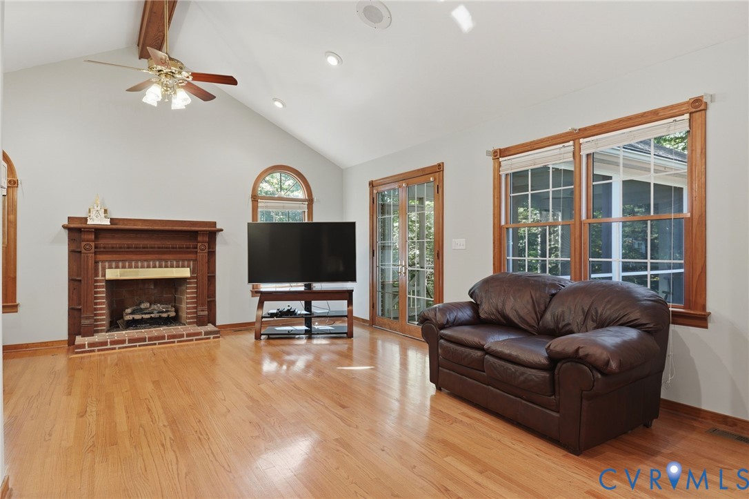 Vaulted ceiling featuring exposed wood beams, hardwood flooring, brick-surround fireplace with wood mantel, arched window, and glass-paned doors