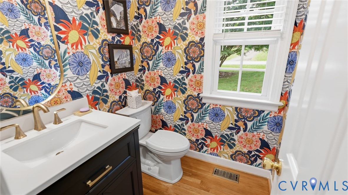 Powder room featuring floral wallpaper, wood-finish flooring, dark-finish cabinetry with a white countertop, and gold-tone fixtures