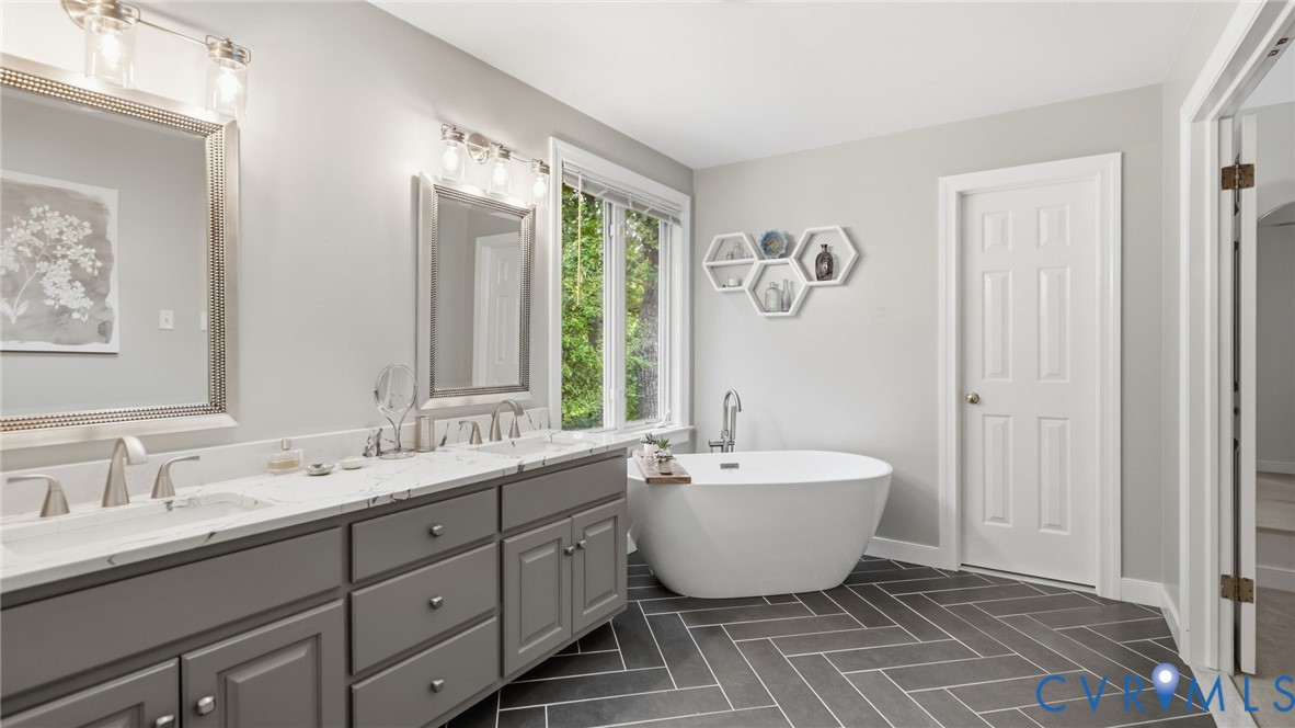 Dual-sink vanity with stone countertop and metallic fixtures, freestanding soaking tub, and herringbone-patterned tile flooring