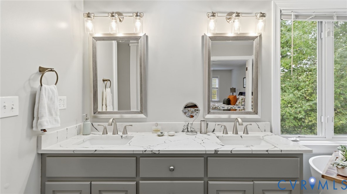 Dual-sink vanity featuring a stone-composite countertop with integrated basins, grey cabinetry, framed wall mirrors, and multi-bulb light fixtures