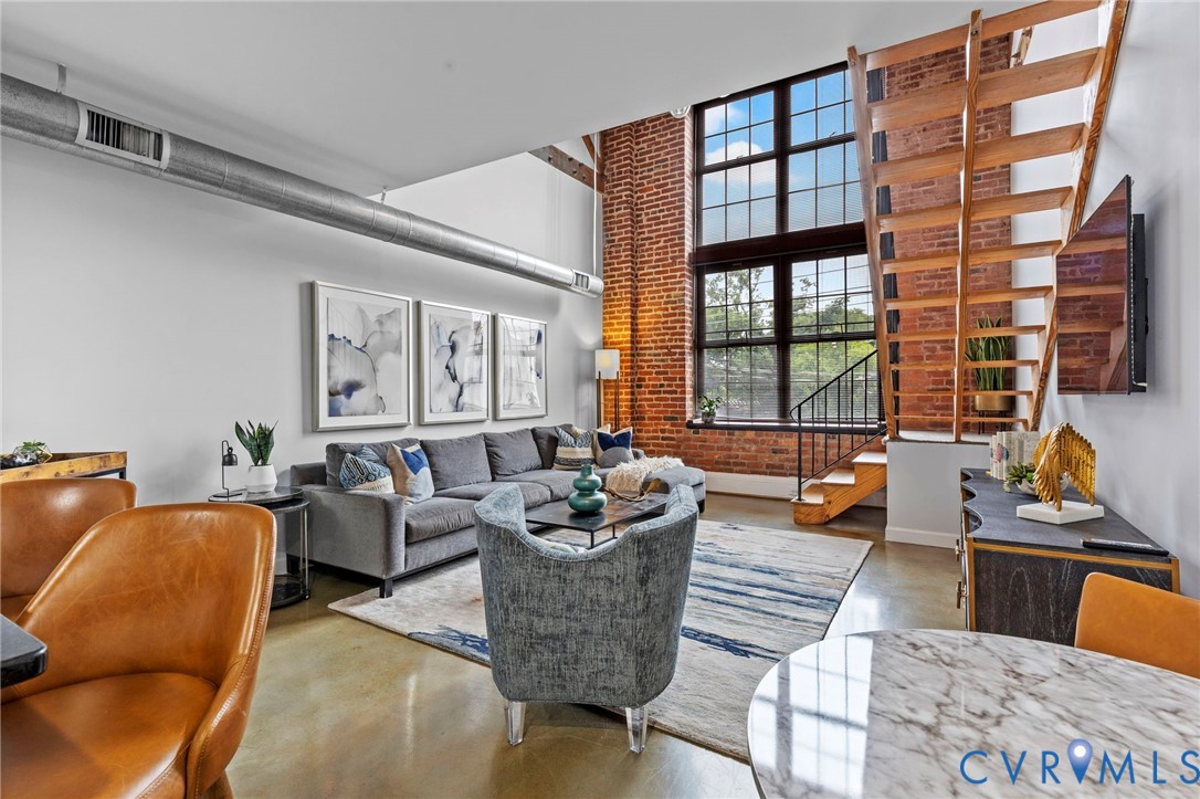 Industrial-style interior featuring soaring ceilings, expansive multi-pane windows, exposed brick accent walls, polished concrete flooring, and an open-tread wooden staircase