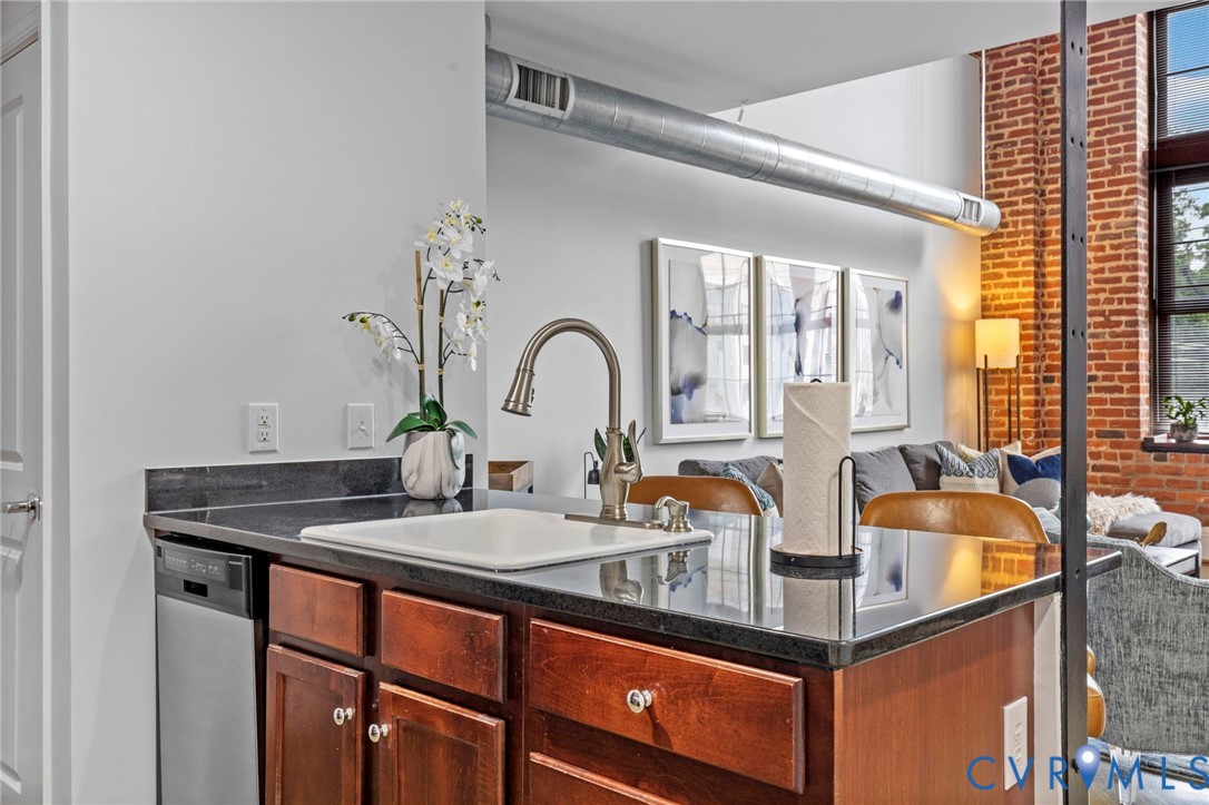 Kitchen island featuring dark stone countertops, an under-mount square sink, gooseneck faucet, warm wood-finish cabinetry, and exposed brick walls