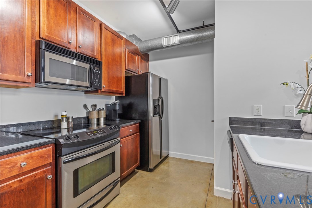Kitchen featuring stained wood cabinetry, dark stone-style countertops, stainless steel appliances, and polished concrete flooring