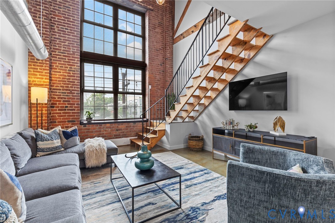 Exposed brick walls, double-height industrial-style windows, open-tread wooden staircase with metal handrail, polished concrete flooring, and exposed ceiling ductwork