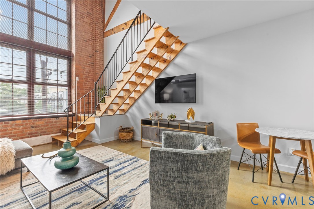 Industrial-style interior featuring floor-to-ceiling multi-pane windows, exposed brick wall, open-riser staircase with metal railing, and concrete flooring