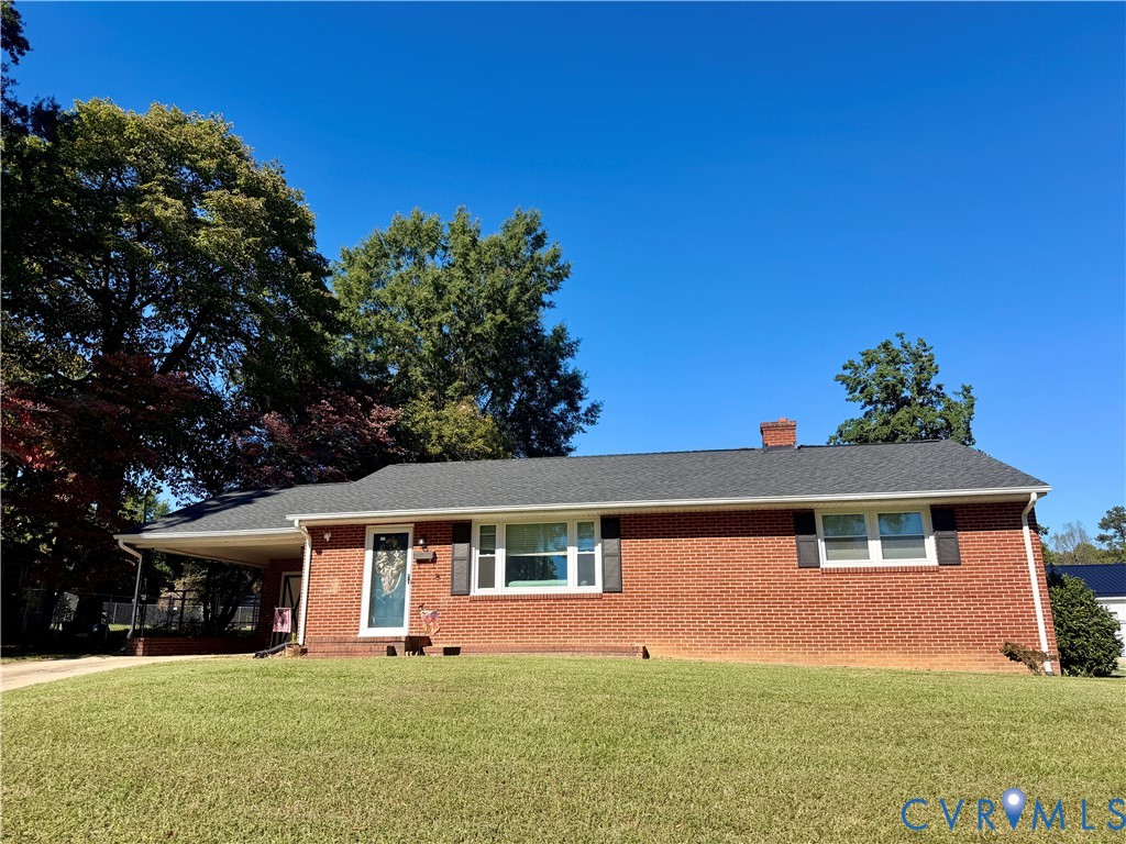 Red brick exterior featuring a covered carport, asphalt shingle roof, dark shutters, and mature trees
