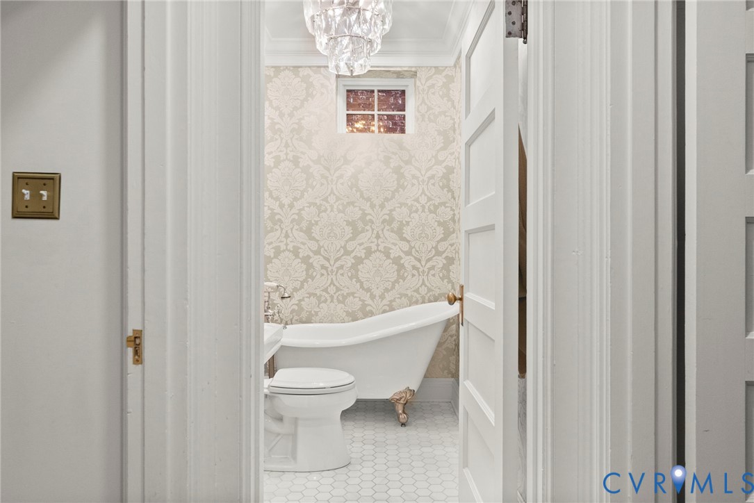 Clawfoot soaking tub with metallic feet, patterned wall covering, crystal chandelier, and hexagonal tile flooring