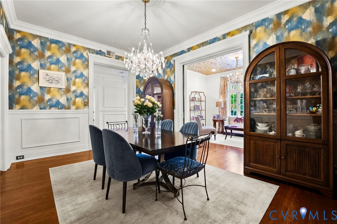 Formal dining space featuring multi-tonal patterned wallpaper, white wainscoting, wood-finish flooring, crown molding, and an ornate crystal chandelier