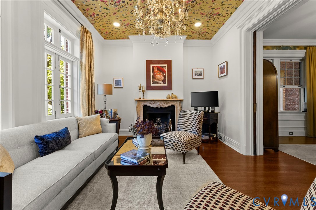Living room featuring an ornate stone fireplace mantel, multi-pane windows, wood-finish flooring, patterned ceiling wallpaper, and a central crystal chandelier