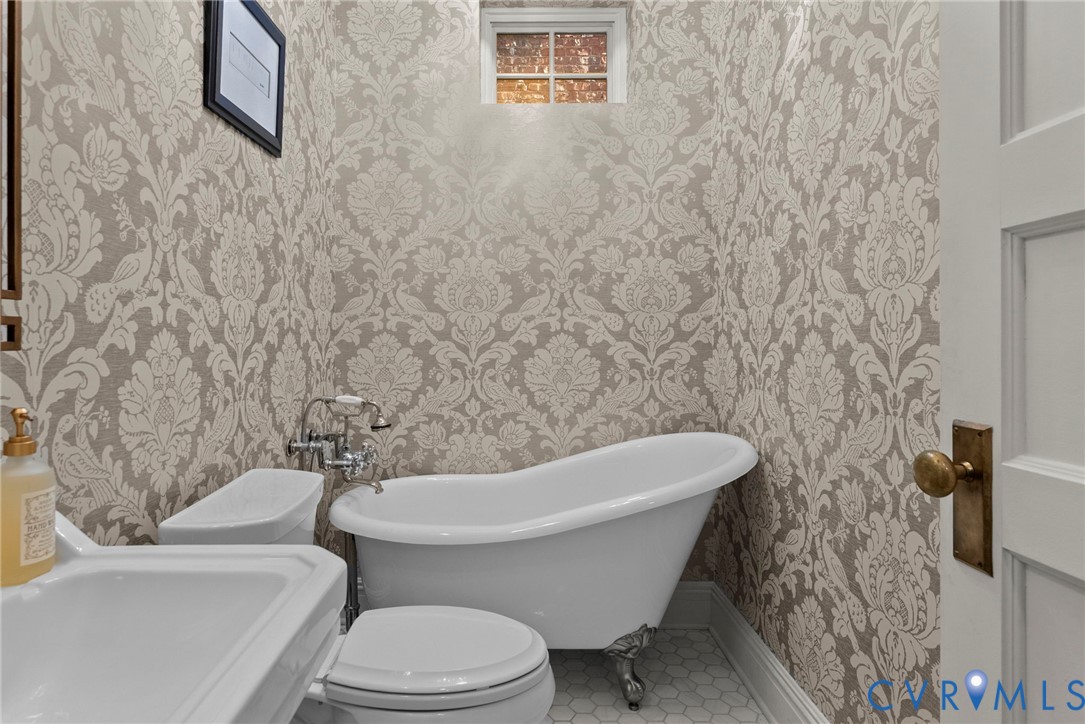 Clawfoot soaking tub with metallic feet, patterned wallpaper, wall-mounted faucet, hexagon tile flooring, and multi-pane window