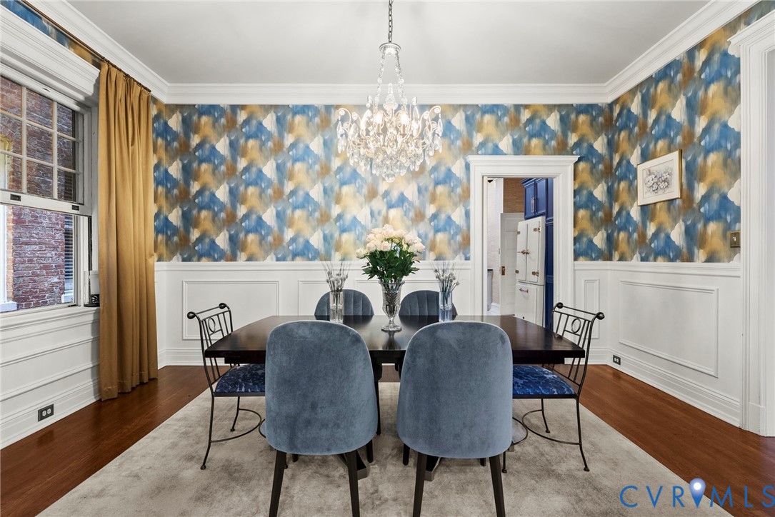 Formal dining space featuring white wainscoting, multi-toned geometric wallpaper, wood-finish flooring, crown molding, and a crystal chandelier