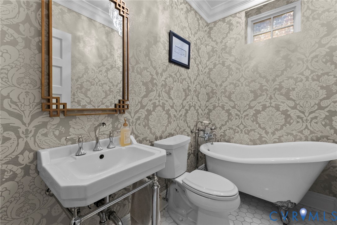 Wall-mounted console sink with exposed plumbing, classic clawfoot soaking tub, patterned wallpaper, wall-mounted mirror with geometric frame, and hexagonal floor tiling