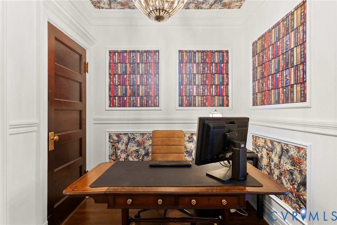 Wood-finish door with brass hardware, wall molding with patterned wallpaper insets, crown molding, ceiling-mounted statement light fixture, and wood-finish flooring