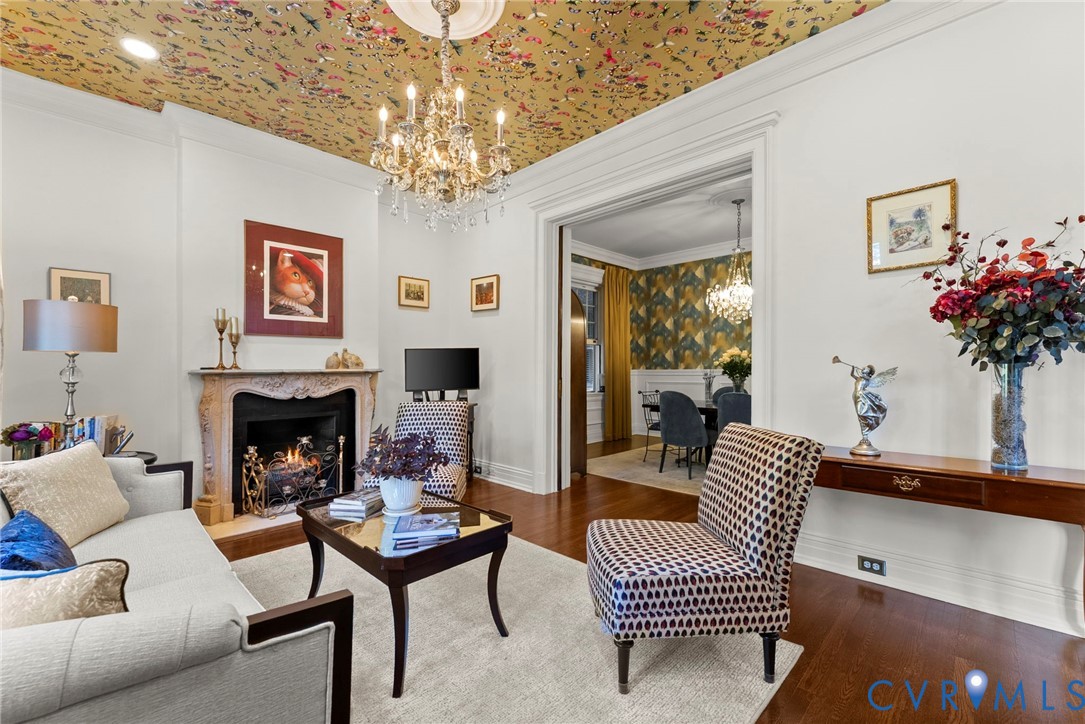 Carved stone fireplace mantle, ceiling medallion, crystal chandelier, floral-patterned ceiling treatment, and wood-finish flooring