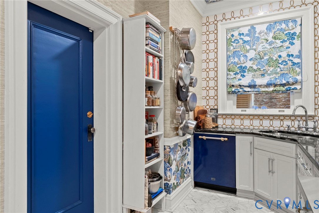 Custom-built narrow shelving unit, patterned wall covering, blue-finished dishwasher, dark stone-finish countertop, and white cabinetry with metal hardware