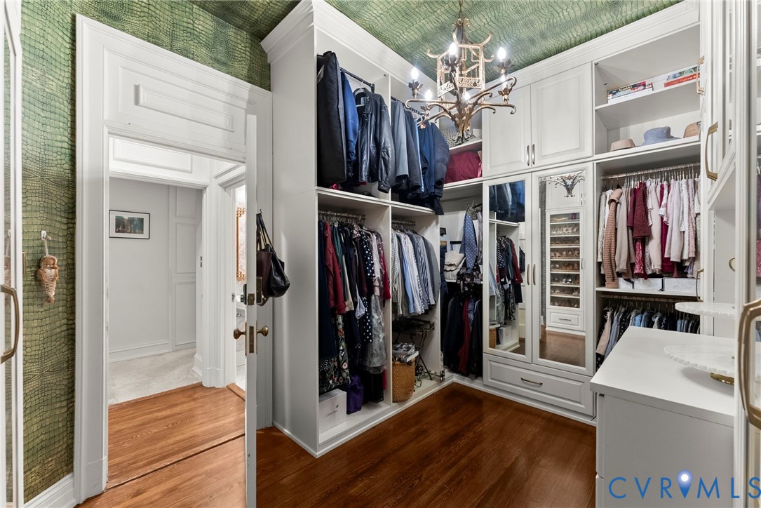 Walk-in closet featuring custom floor-to-ceiling white cabinetry, mirrored wardrobe doors, integrated shelving, hardwood flooring, and a decorative chandelier