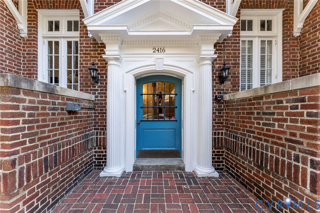 Red brick exterior featuring a classical pedimented entryway, fluted columns, a blue wood-panel door with an iron grille, and a brick-paved stoop