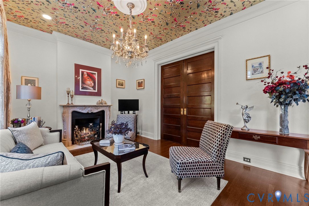 Interior space featuring wood-finish flooring, a decorative fireplace with carved stone surround, a central crystal chandelier with ceiling medallion, patterned wallpapered ceiling, and solid wood double doors
