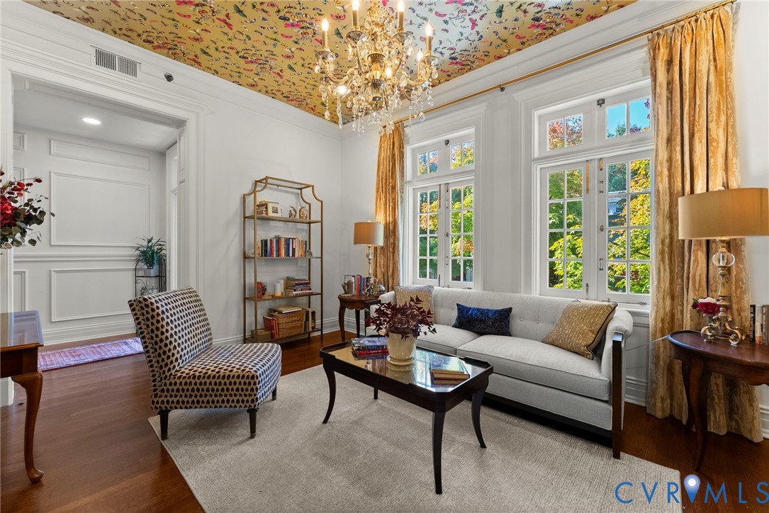 Living area featuring wood-finish flooring, decorative ceiling paper, multi-pane windows, and an ornate crystal chandelier