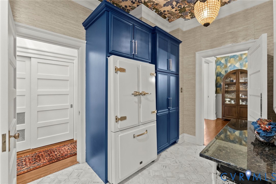 Custom floor-to-ceiling blue cabinetry with integrated paneled refrigerator, patterned ceiling wallpaper, natural fiber wall covering, and stone tile flooring