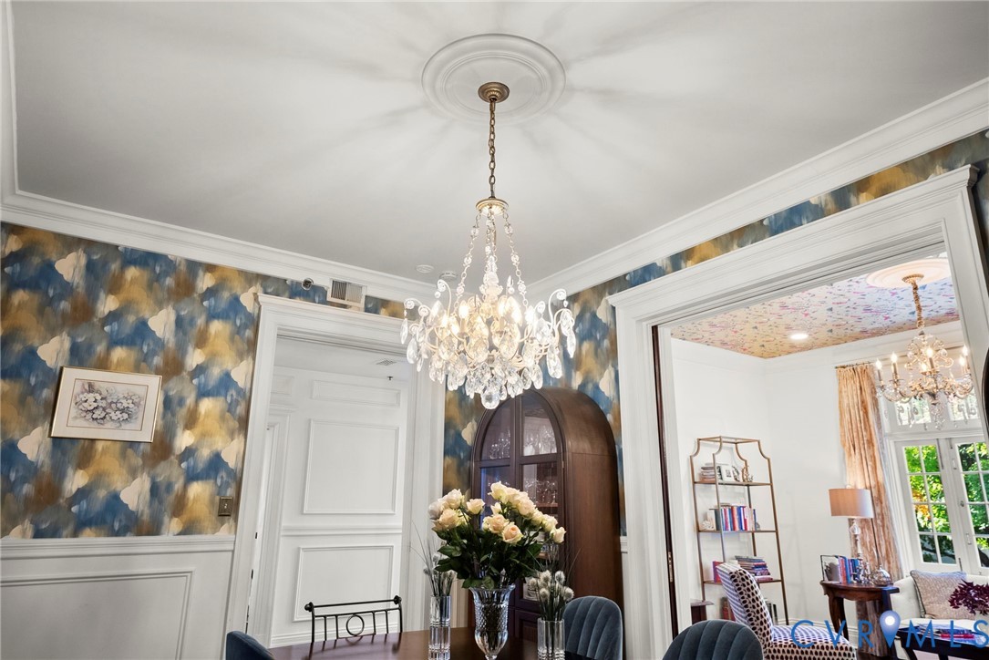 Ornate crystal chandelier with central ceiling medallion, extensive crown molding, decorative wainscoting, and bold patterned wallpaper